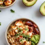 Colorful Southwest Chicken Burrito Bowls featuring seasoned grilled chicken, vibrant red and green bell peppers, black beans, corn, crispy tortilla strips, all arranged in a white ceramic bowl. The dish is topped with fresh cilantro, shredded cheese, and a drizzle of creamy sauce, styled with fresh lime wedges on a rustic wooden table with a bright, inviting background.