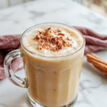 A beautifully styled clear glass mug filled with a rich, creamy vanilla cinnamon latte topped with a swirl of whipped cream, cinnamon dusting, and vanilla bean pod garnish. The latte displays a warm caramel hue, with layered textures from the foam and coffee, set against a rustic wooden table with cinnamon sticks and vanilla beans nearby, styled for a cozy, inviting atmosphere.